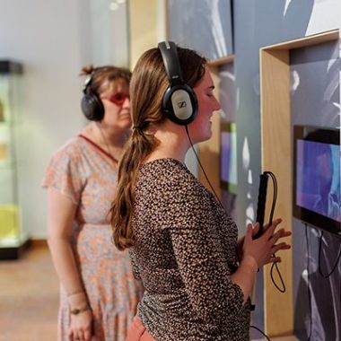 In het Gasthuismuseum Geel hangen drie schermen aan de muur, waaraan koptelefoons verbonden zijn via kabels. Een blinde en een slechtziende vrouw hebben allebei zo’n koptelefoon op en beluisteren het filmpje dat afspeelt op de schermen. 