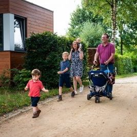 Een gezin van zes wandelt over een zandpad in Camping Houtum. Naast het pad staat een van de autismevriendelijke, houten bungalows van het domein. De vader, gekleed in een paarse polo, duwt een kinderwagen terwijl de moeder een klein kind op haar schouders draagt. Voor hen loopt een kleine jongen in een rood T-shirt vrolijk vooruit, terwijl een iets oudere jongen in een blauw shirt naast hen wandelt.