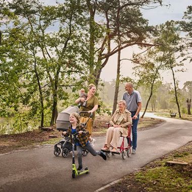 Een moeder wandelt met haar twee kinderen op een verharde ondergrond tussen de bomen aan het water. Eén van haar kinderen is nog een baby en houdt ze in haar rechterarm, terwijl ze met haar linkerarm een buggy duwt. Het oudere zoontje gaat snel vooruit op zijn step. Naast dit drietal wandelt een ouder koppel: de vrouw zit in een rolstoel, die wordt voortgeduwd door haar echtgenoot. 