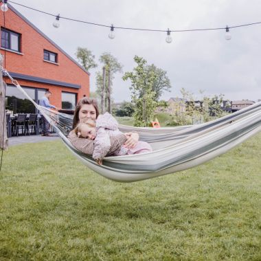 Bij vakantiewoning Den Akker in Mol ligt een moeder samen met haar jonge dochter in een stoffen hangmat. Ze genieten van de rust op het grasveldje in de landelijke omgeving. Op de achtergrond barbecuet de vader op het betegelde terras. Naast hem staat een lange tuintafel met lege stoelen.