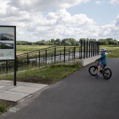 Een jong kind staat met de fiets bij een informatiebord, geplaatst naast de ontwateringssluis van het overstromingsgebied Polder van Lier.