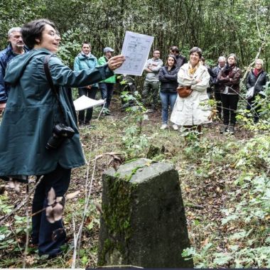 Een persoon staat naast een oude grenssteen en spreekt tot een groep geïnteresseerde wandelaars.