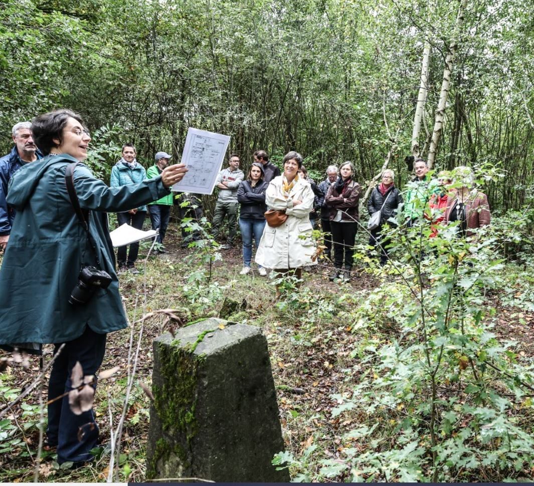 Een persoon staat naast een oude grenssteen en spreekt tot een groep geïnteresseerde wandelaars.