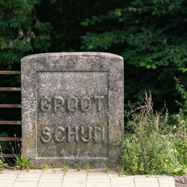 Een oude naamsteen draagt de naam 'Groot Schijn'.  De staan staat aan de rand van de stoep, met aan de linkerzijde een roestige leuning. Achter steen en leuning ligt een diepte waar bomen staan.