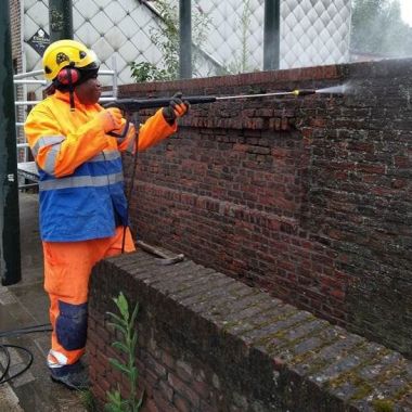 Een persoon in een werkpak en met helm spuit een bakstenen muurtje schoon met een hogedrukspuit.