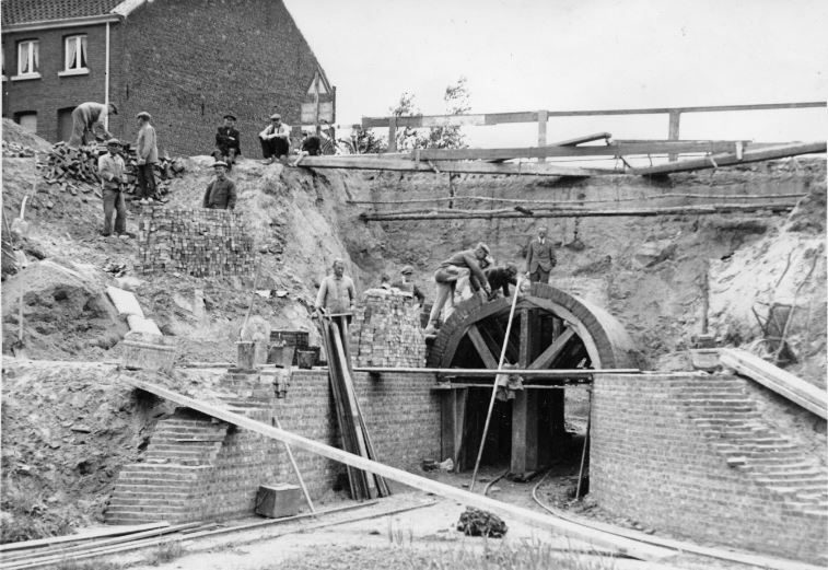 Zwart-wit foto van een tunnel in opbouw. Enkele arbeiders staan bovenop de tunnelboog en leggen bakstenen. Op de helling naast de tunnel staan nog enkele arbeiders bij stapels bakstenen. Vanop  de hogergelegen straat kijken enkele mensen gehurkt toe. De straat is bovenaan afgezet met een houten hekwerk. Op de voorgrond liggen planken en ander bouwmateriaal. 