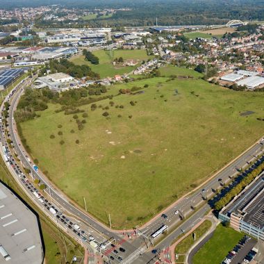 Een groene grasvlakte is aan twee zijden omzoomd met drukke verkeerswegen. Rechts vooraan liggen de gebouwen van het Wijnegem Shopping Center. 