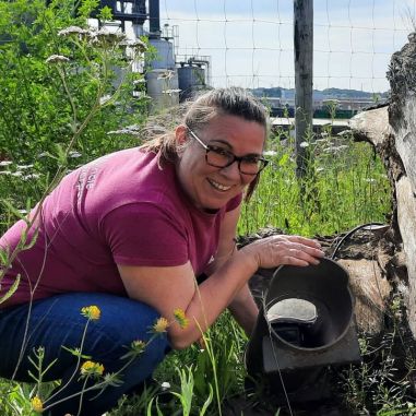 Een provinciemedewerker zit gehurkt naast een wildcamera in een buis. Ernaast ligt een grote boomstronk in het gras en tussen de bloemen.  Achter hen is een afsluiting te zien.