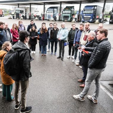 Een groep van een dertigtal mensen staat in een kring buiten op het asfalt. Achter hen is een rij vrachtwagens zichtbaar die brandstof tanken bij een benzinestation.