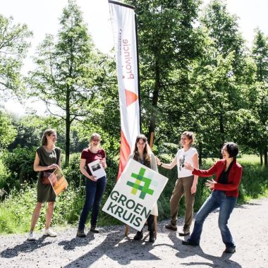 Vijf personen staan op een landweg voor een groepje bomen. Achter hen staat een strandvlag met het logo van de provincie Antwerpen. De middelste persoon draagt een groot bord met het logo van Groen Kruis.