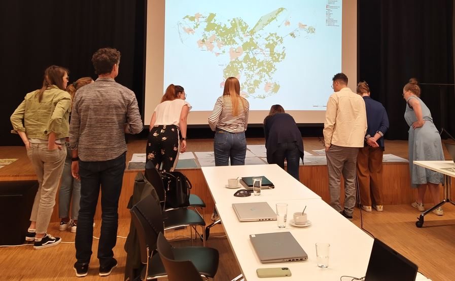 Een groep personen staat in een zaal gebogen over kaarten en tabellen op tafels. Voor hen is een grote projectie zichtbaar van de warmtezoneringskaart voor Energielandschap Noordertuin.
