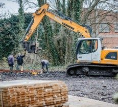 Een graafmachine staat voor enkele bomen en een loods. Arbeiders bevestigen een hijstouw van de graafarm rond enkele houten planken. Op de voorgrond staat een pallet met nog meer houten planken.