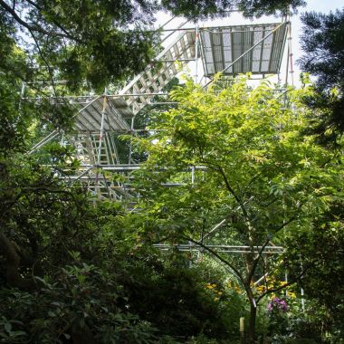 Doorkijk tussen bomen, struiken en groene borders naar een hoge stelling met uitkijkplatformen op de achtergrond, in Arboretum Kalmthout.