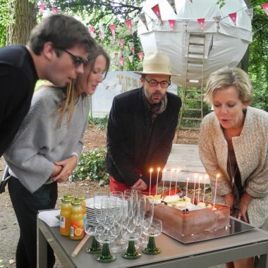 Een jong koppeltje en een man en vrouw blazen tien kaarsjes op een taart uit, op de achtergrond een bolvormige tent in een park, er hangen vlaggenslingers op. Viering 10 jaar Terra Nova in Arboretum Kalmthout.