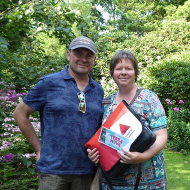 Een man en een vrouw poseren lachend naast bloeiende borders in de tuin van Arboretum Kalmthout. De vrouw houdt een schoudertas vast en toont een ticket waar Terra Nova op staat.