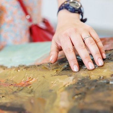 Een close-up van een hand die streelt over een schilderij in het Jakob Smits-museum. Het schilderij ligt horizontaal en bestaat uit dikke lagen verf en bruuske penseelstreken in donkere kleuren. De hand bevoelt het reliëf van de verf. 