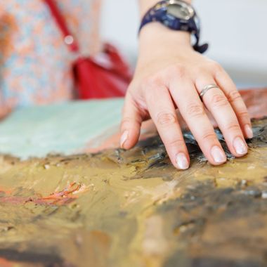 Een close-up van een hand die streelt over een schilderij in het Jakob Smits-museum. Het schilderij ligt horizontaal en bestaat uit dikke lagen verf en bruuske penseelstreken in donkere kleuren. De hand bevoelt het reliëf van de verf.