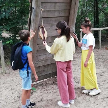 Drie kinderen ontdekken in provinciaal domein Hoge Mouw het bosspel bij een houten speeltuig op een zanderige ondergrond. De jongen leunt tegen het speeltuig en draagt de rugzak met de spelattributen, terwijl het meisje rechts het gps-toestel bedient. Een ander meisje staat in het midden en houdt een aanwijzing van het bosspel vast in haar linkerhand.