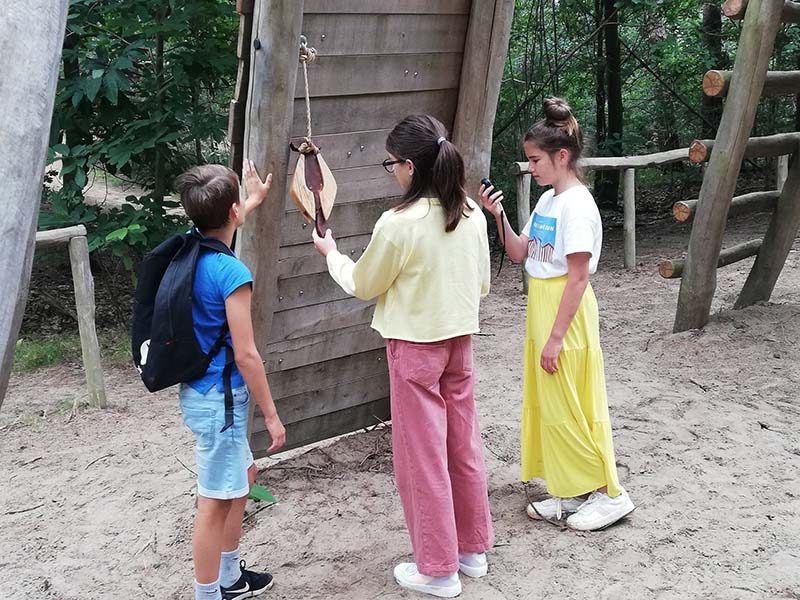 Drie kinderen ontdekken in provinciaal domein Hoge Mouw het bosspel bij een houten speeltuig op een zanderige ondergrond. De jongen leunt tegen het speeltuig en draagt de rugzak met de spelattributen, terwijl het meisje rechts het gps-toestel bedient. Een ander meisje staat in het midden en houdt een aanwijzing van het bosspel vast in haar linkerhand.