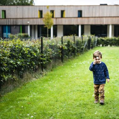 In de afgeschermde tuin van vakantieverblijf Casa Ametza van vzw Bielebale in Brasschaat bevindt zich een kleine kleuter. Hij houdt een witte veldbloem omhoog terwijl hij over het gras wandelt. 