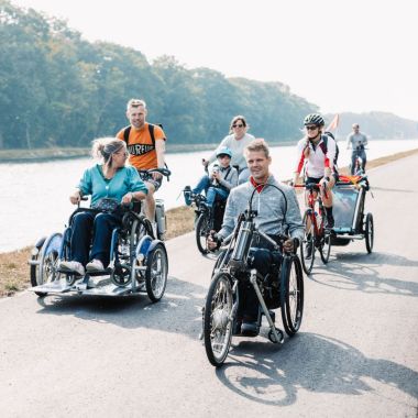 Een groepje van zeven personen fietst op het jaagpad langs het kanaal Dessel Schoten in Mol. Ze zitten elk op een toegankelijke fiets: de eerste man beweegt zich voort met een handbike, een vrouw zit op een rolstoelfiets die wordt bestuurd door haar echtgenoot. Daarachter fietst een vrouw met haar zoon in een semi-ligfiets tandem. Een andere vrouw die achterop fietst, draagt een helm en heeft een fietskar achter aan haar fiets bevestigd. De zon schijnt.