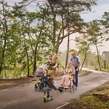 Een moeder wandelt met haar twee kinderen op een verharde ondergrond tussen de bomen aan het water. Eén van haar kinderen is nog een baby en houdt ze in haar rechterarm, terwijl ze met haar linkerarm een buggy duwt. Het oudere zoontje gaat snel vooruit op zijn step. Naast dit drietal wandelt een ouder koppel: de vrouw zit in een rolstoel, die wordt voortgeduwd door haar echtgenoot. 