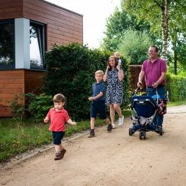 Een gezin van zes wandelt over een zandpad in Camping Houtum. Naast het pad staat een van de autismevriendelijke, houten bungalows van het domein. De vader, gekleed in een paarse polo, duwt een kinderwagen terwijl de moeder een klein kind op haar schouders draagt. Voor hen loopt een kleine jongen in een rood T-shirt vrolijk vooruit, terwijl een iets oudere jongen in een blauw shirt naast hen wandelt.