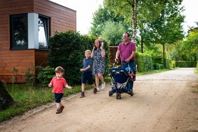 Een gezin van zes wandelt over een zandpad in Camping Houtum. Naast het pad staat een van de autismevriendelijke, houten bungalows van het domein. De vader, gekleed in een paarse polo, duwt een kinderwagen terwijl de moeder een klein kind op haar schouders draagt. Voor hen loopt een kleine jongen in een rood T-shirt vrolijk vooruit, terwijl een iets oudere jongen in een blauw shirt naast hen wandelt.