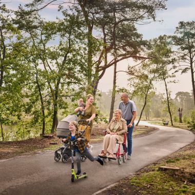 Een moeder wandelt met haar twee kinderen op een verharde ondergrond tussen de bomen aan het water. Eén van haar kinderen is nog een baby en houdt ze in haar rechterarm, terwijl ze met haar linkerarm een buggy duwt. Het oudere zoontje gaat snel vooruit op zijn step. Naast dit drietal wandelt een ouder koppel: de vrouw zit in een rolstoel, die wordt voortgeduwd door haar echtgenoot. 