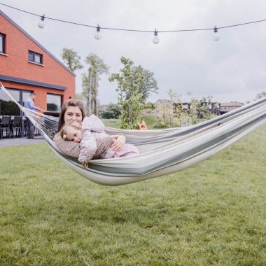 Bij vakantiewoning Den Akker in Mol ligt een moeder samen met haar jonge dochter in een stoffen hangmat. Ze genieten van de rust op het grasveldje in de landelijke omgeving. Op de achtergrond barbecuet de vader op het betegelde terras. Naast hem staat een lange tuintafel met lege stoelen.